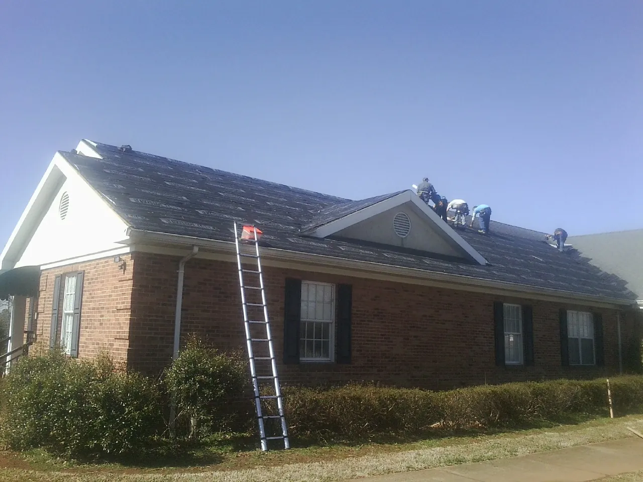 Crew tearing shingles off a brick ranch roof with a ladder set — Metal Roof Repair work by our crew in Elwell