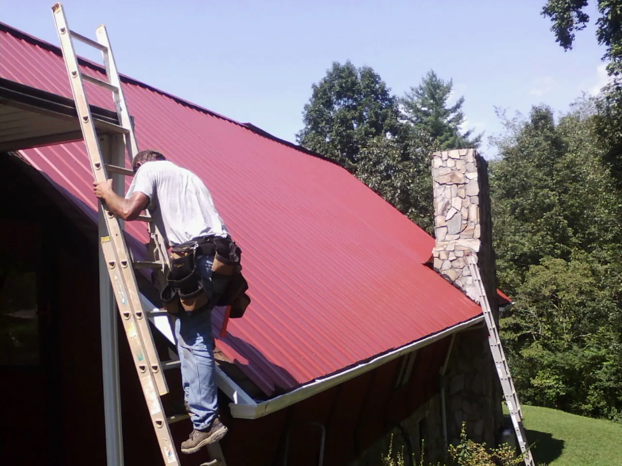 Roofer on a ladder beside a red metal roof and stone chimney — Soffit & Fascia Repair by Roofing Brothers in Elwell